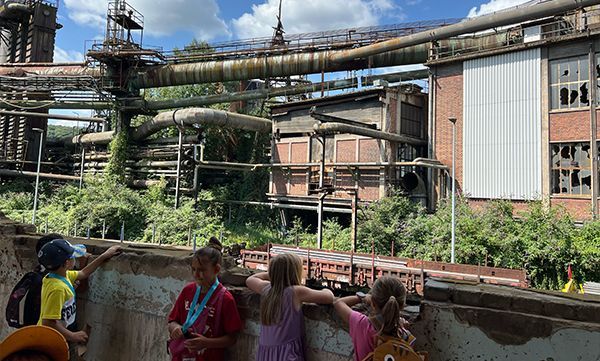 Kinder betrachten ein verlassenes Industriegebäude mit wucherndem Bewuchs; sonniger Tag.