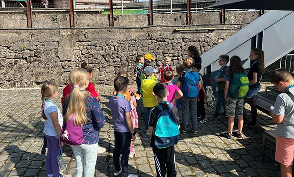 Eine Gruppe Kinder steht im Freien und hört jemandem zu. Im Hintergrund eine Steinmauer. Sonniger Tag.