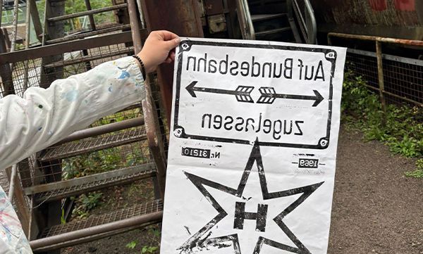 Eine Person hält ein Schild mit der Aufschrift „Auf Bundesbahn zugelassen“ und Pfeilen. Auf dem Schild befindet sich ein Stern mit einem „H“ darin.