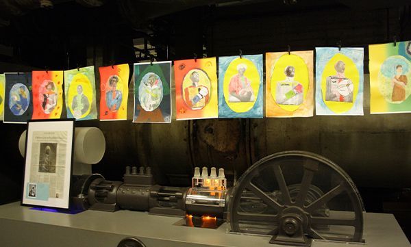 Ausstellung von Kunstwerken und Maschinen. Farbenfrohe Porträts hängen über mechanischen Teilen.