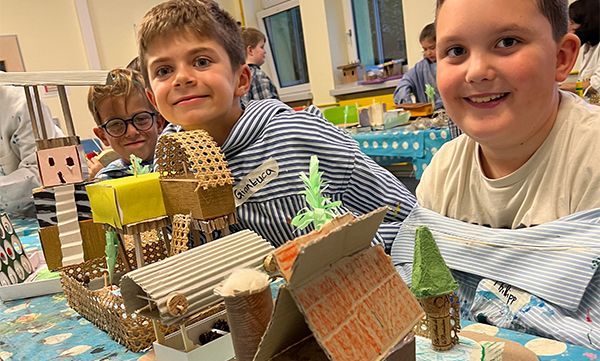 Lächelnde Kinder mit ihren Bastelprojekten auf einem Tisch, darunter Gebäude aus Pappe und anderen Materialien.