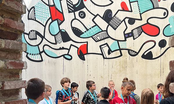 Kinder betrachten ein farbenfrohes abstraktes Wandgemälde an einer Betonwand; links davon eine Backsteinmauer.