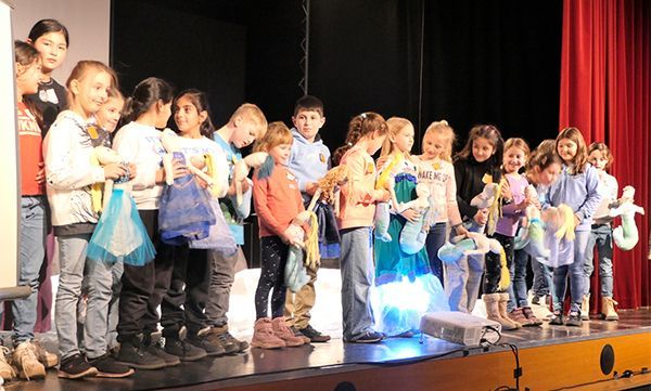 Kinder mit Spielzeug und Geschenktüten auf der Bühne führen ein Konzert auf. Im Hintergrund rote Vorhänge.