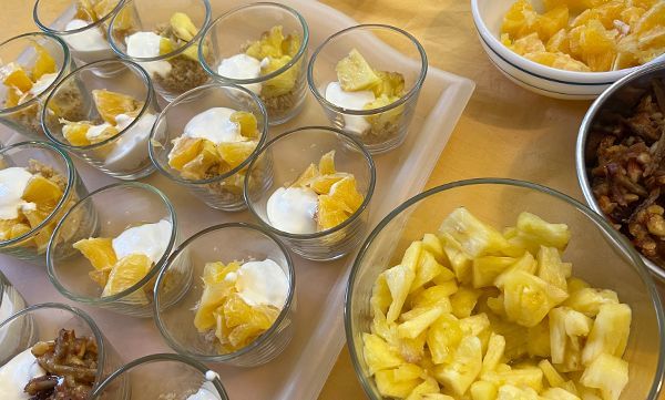 Dessertgläser mit geschichteten Früchten, Joghurt und Nüssen. Schüsseln mit geschnittenem Obst und Nüssen stehen daneben.
