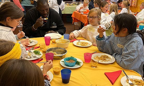 Menschen essen an einem Tisch mit Tellern voller Speisen. Ein Mann und Kinder sitzen um die gelbe Tischdecke versammelt.