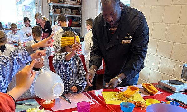 Ein Koch führt ein Kind mit verbundenen Augen an einem Kochtisch mit Zutaten. Im Hintergrund sind weitere Kinder und ein Erwachsener zu sehen.