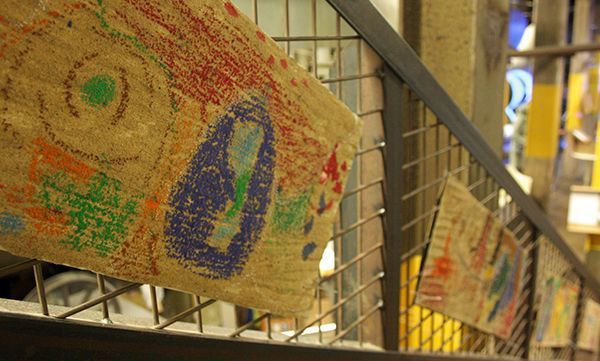 Bunte Buntstiftzeichnungen auf Jute, die an einem Metallgeländer in einem Gebäude angebracht sind.