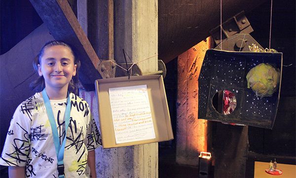 Ein Mädchen steht neben einer Kunstausstellung, die einen gemalten Nachthimmel, einen Planeten und eine handgeschriebene Notiz in einem Papprahmen zeigt.