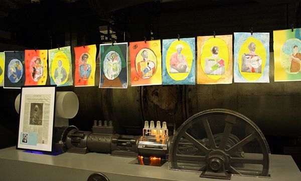 Eine Kunstausstellung mit farbenfrohen Porträts hängt über Industriemaschinen.