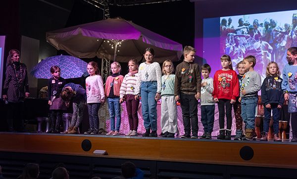 Kinder auf einer Bühne mit einer Frau und einem Regenschirm als Dekoration. Sie scheinen an einer Aufführung oder Veranstaltung teilzunehmen.