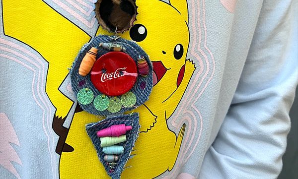 Eine Person, die ein hellblaues Pikachu-Sweatshirt und eine handgefertigte Jeanskette mit einem Coca-Cola-Knopf und Buntstiftdetails trägt.