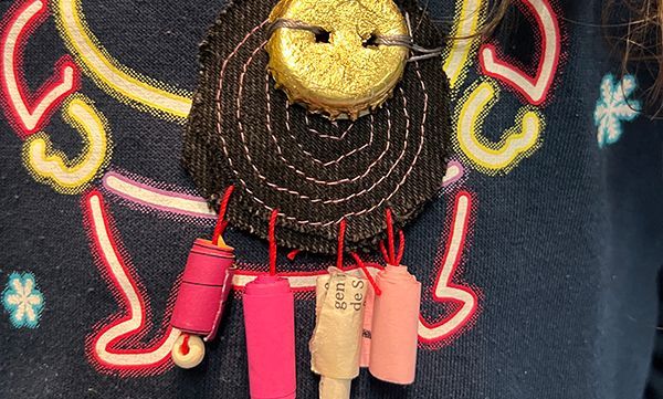 Nahaufnahme einer handgefertigten Halskette aus blauem Jeansstoff. Sie besteht aus einem glitzernden Flaschenverschluss, schwarzem Stoff und bunten, hängenden Papierrollen.