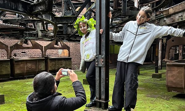 Drei Personen posieren in einer Industrieanlage; eine macht ein Foto, während sich zwei an eine dunkle Säule lehnen. Eine Person trägt eine grüne Blätterkrone.