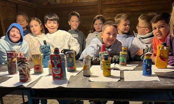 Kinder sitzen an einem Tisch mit bunten, totemartigen Skulpturen. Sie lächeln in einer Werkstattatmosphäre.