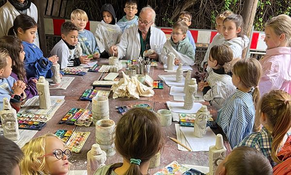 Kinder und Kursleiter an einem Tisch beim Bemalen von Keramikskulpturen. Draußen, leuchtende Farben, konzentrierte Blicke.
