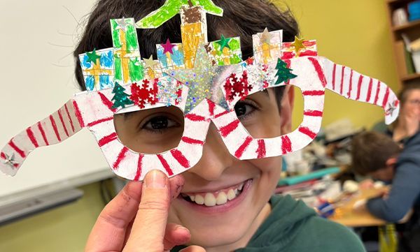 Ein lächelnder Junge trägt eine bunte, weihnachtlich verzierte Papierbrille. Die Bügel sind rot-weiß gestreift, der Brillenkopf mit stadtähnlichen Dekorationen verziert.