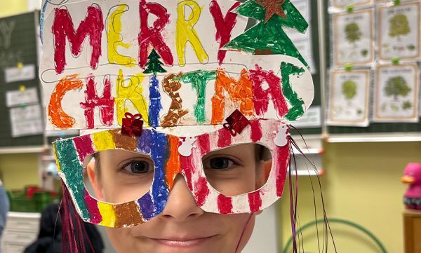 Junge mit bunter, selbstgemachter „Frohe Weihnachten“-Brille und Stirnband in einem Klassenzimmer.