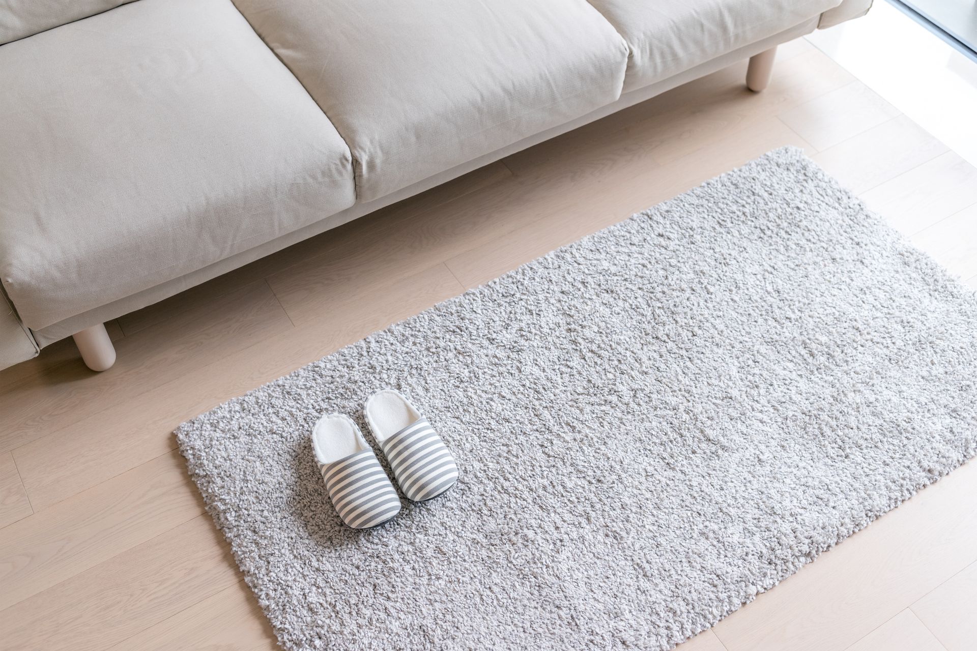 A pair of slippers on a grey carpet on a wooden floor in front of a lighter grey couch.