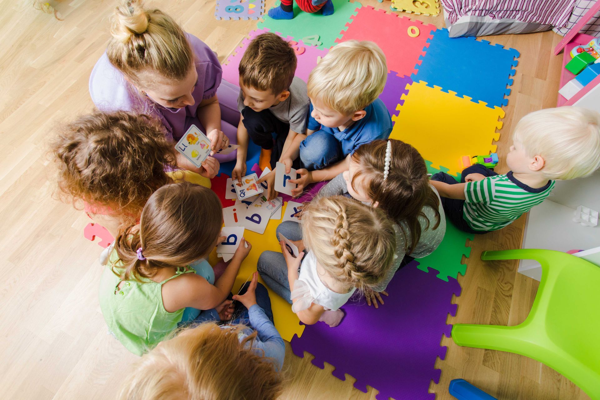 Des enfants, réunis autour d'une table, manipulent des blocs et des papiers colorés sur des tapis en mousse dans une salle de classe.