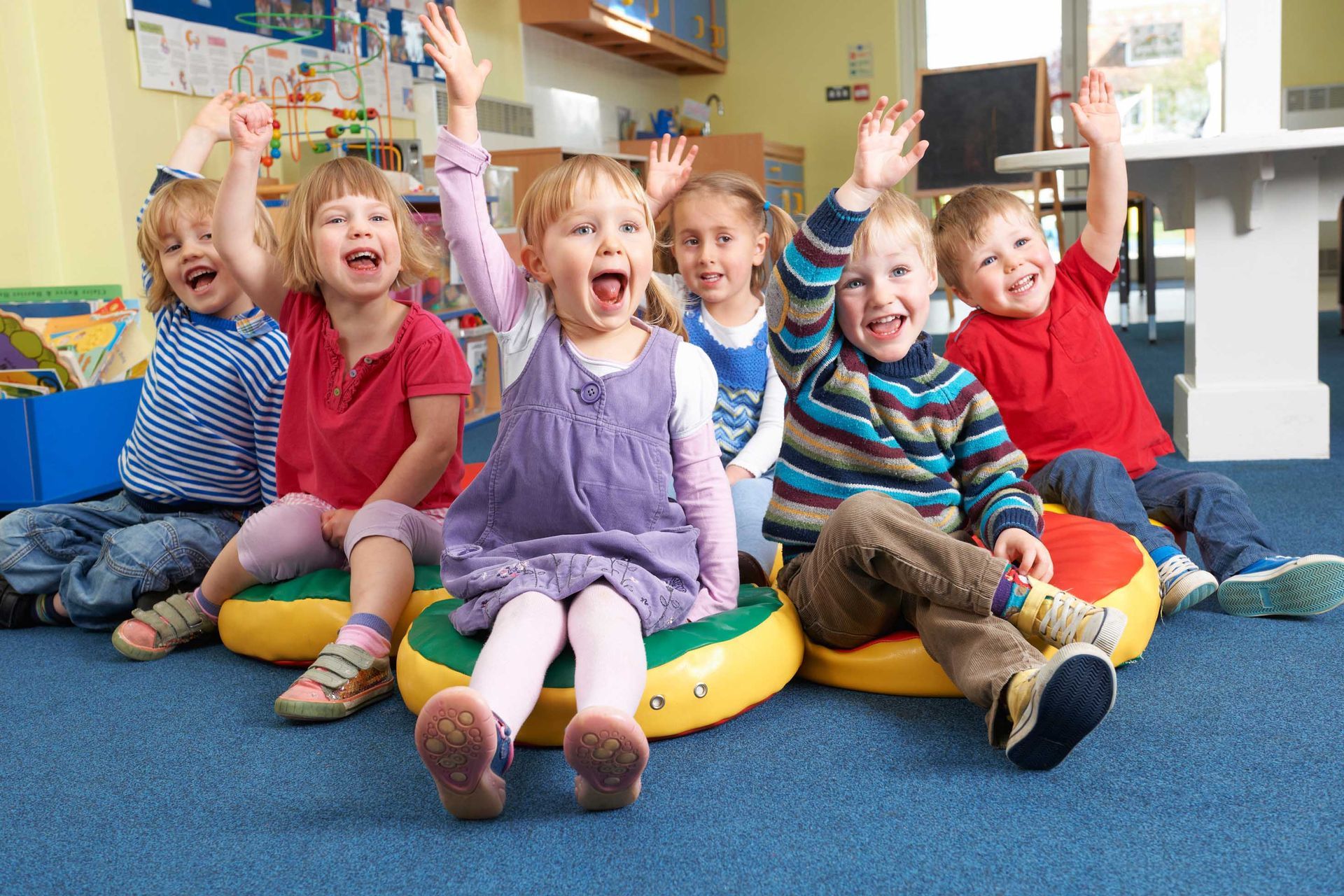 Des enfants assis sur un tapis bleu dans une salle de classe, souriants et levant les mains
