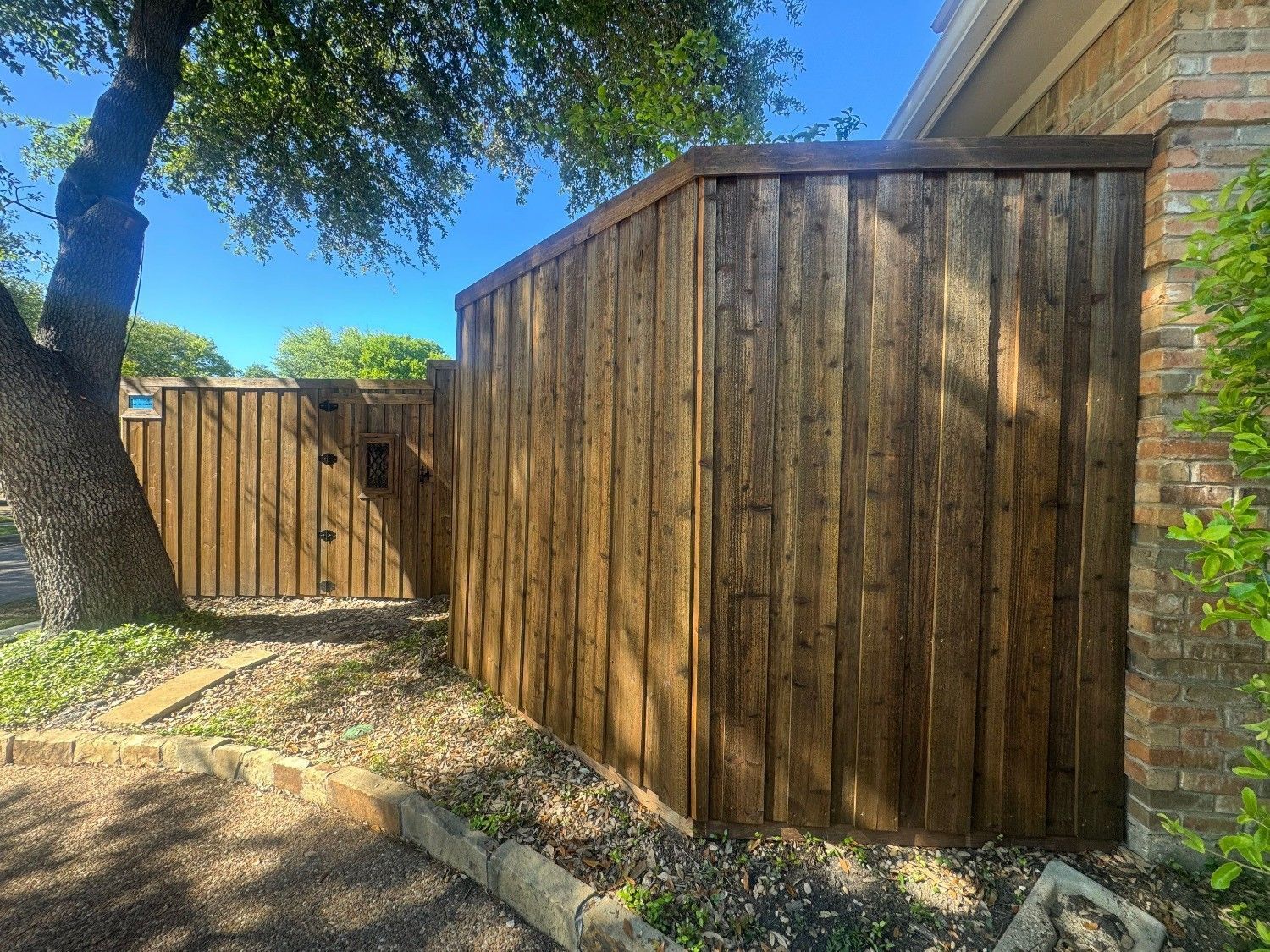 Wooden privacy fence along a stone-edged walkway beside a brick house and trees