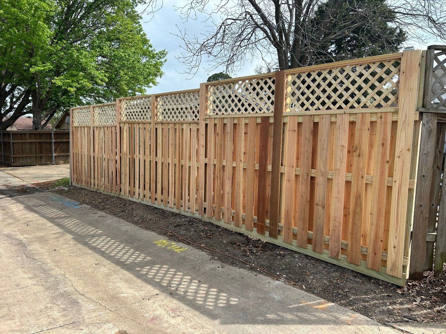 Wooden privacy fence with lattice top lining a paved driveway beside trees.