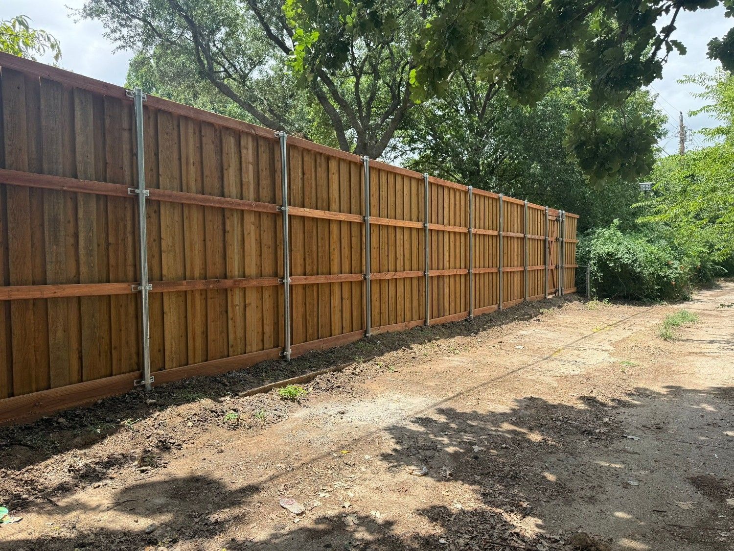 Long wooden privacy fence along a dirt yard with trees and greenery behind it