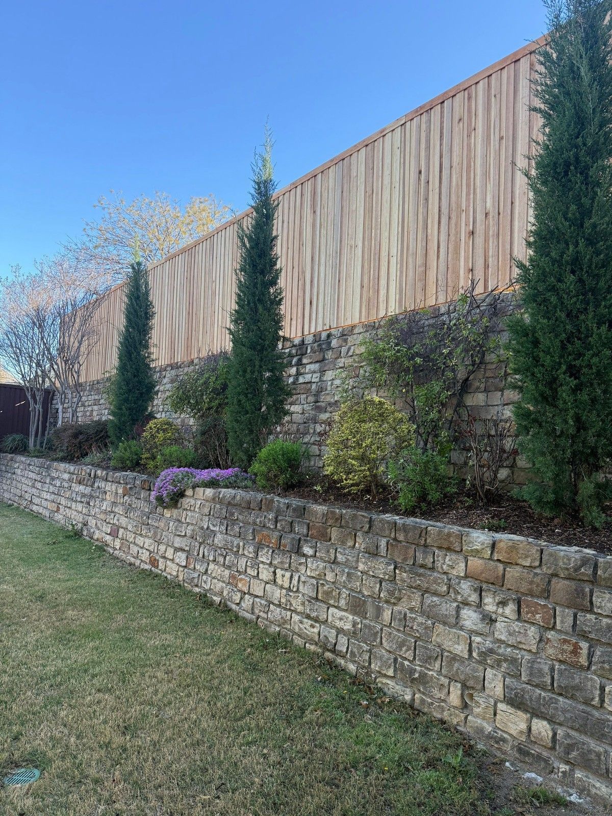 Stone retaining wall with shrubs and flowers below a tall wooden privacy fence along a grassy yard