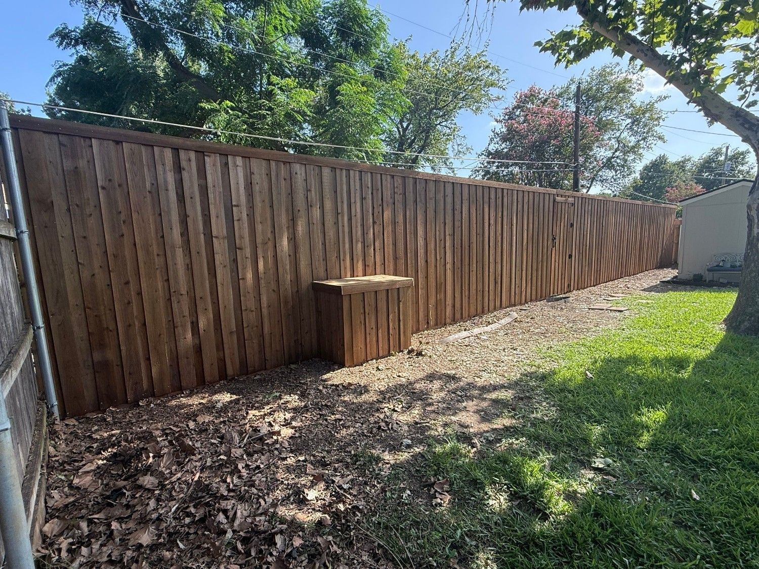 Wooden fence along a backyard with mulch, a small round table, and green grass under sunny trees