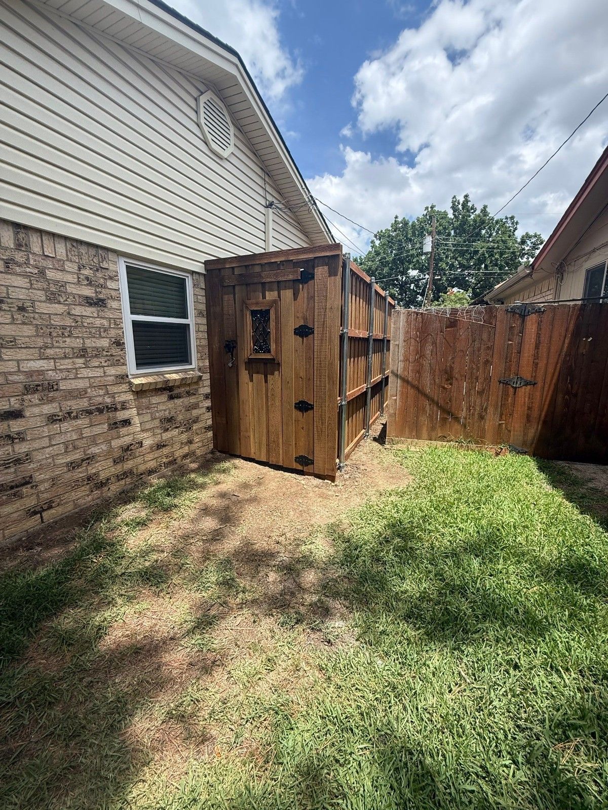 Backyard with wooden shed beside a house, fenced yard, grass, and partly cloudy sky.