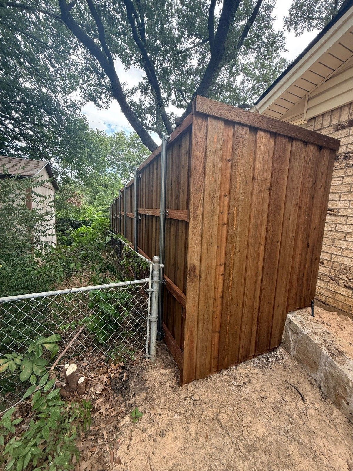 Wooden outdoor storage shed beside a brick house and chain-link fence under trees