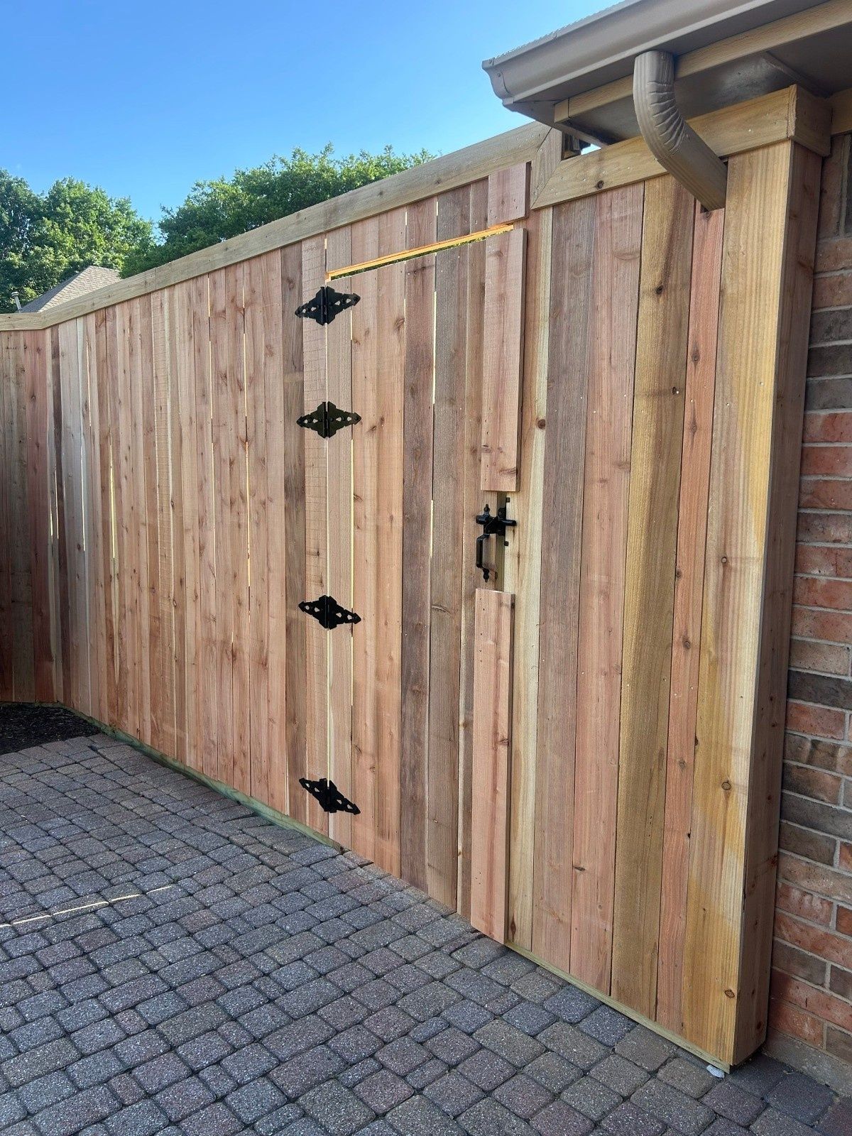 Wooden driveway gate with black hinges beside a brick wall and cobblestone pavement.