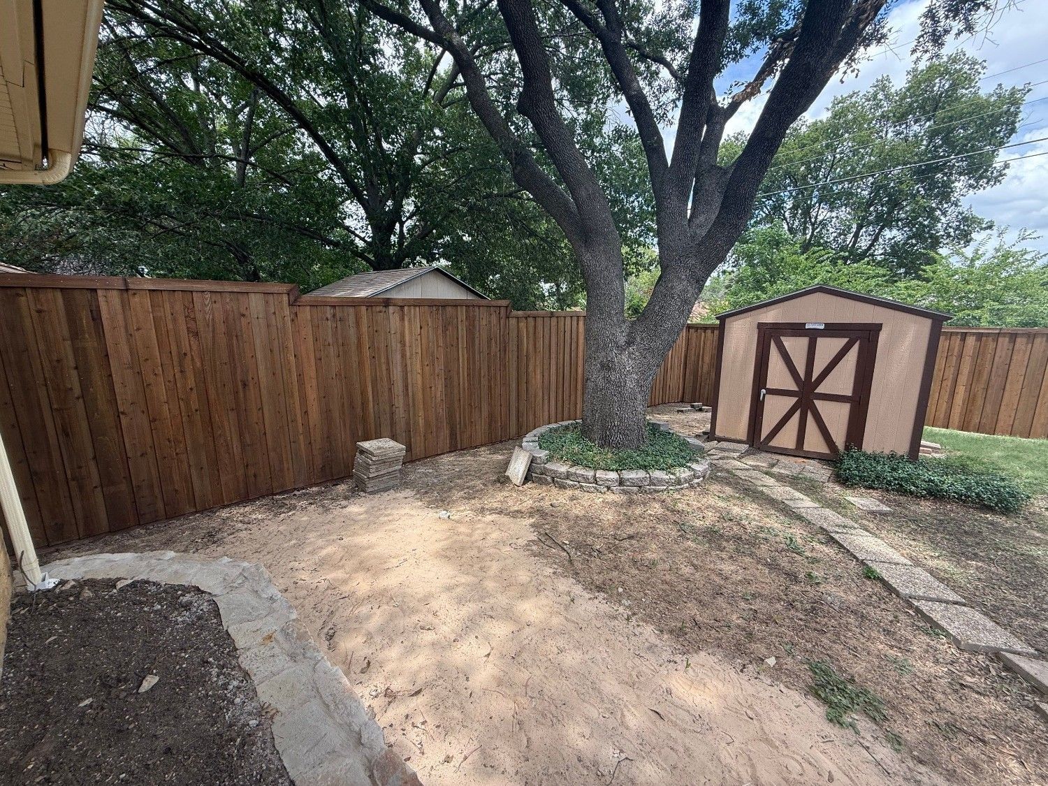 Backyard with large tree, wooden fence, shed, and dirt ground beside a concrete patio