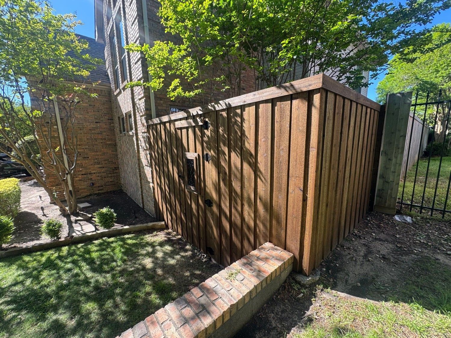 Backyard wooden privacy fence beside a brick house and stone path, with green lawn and trees nearby