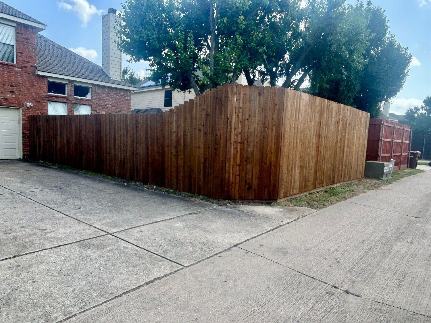 Corner lot with a tall wooden fence beside a driveway, brick house, and trees in the background.