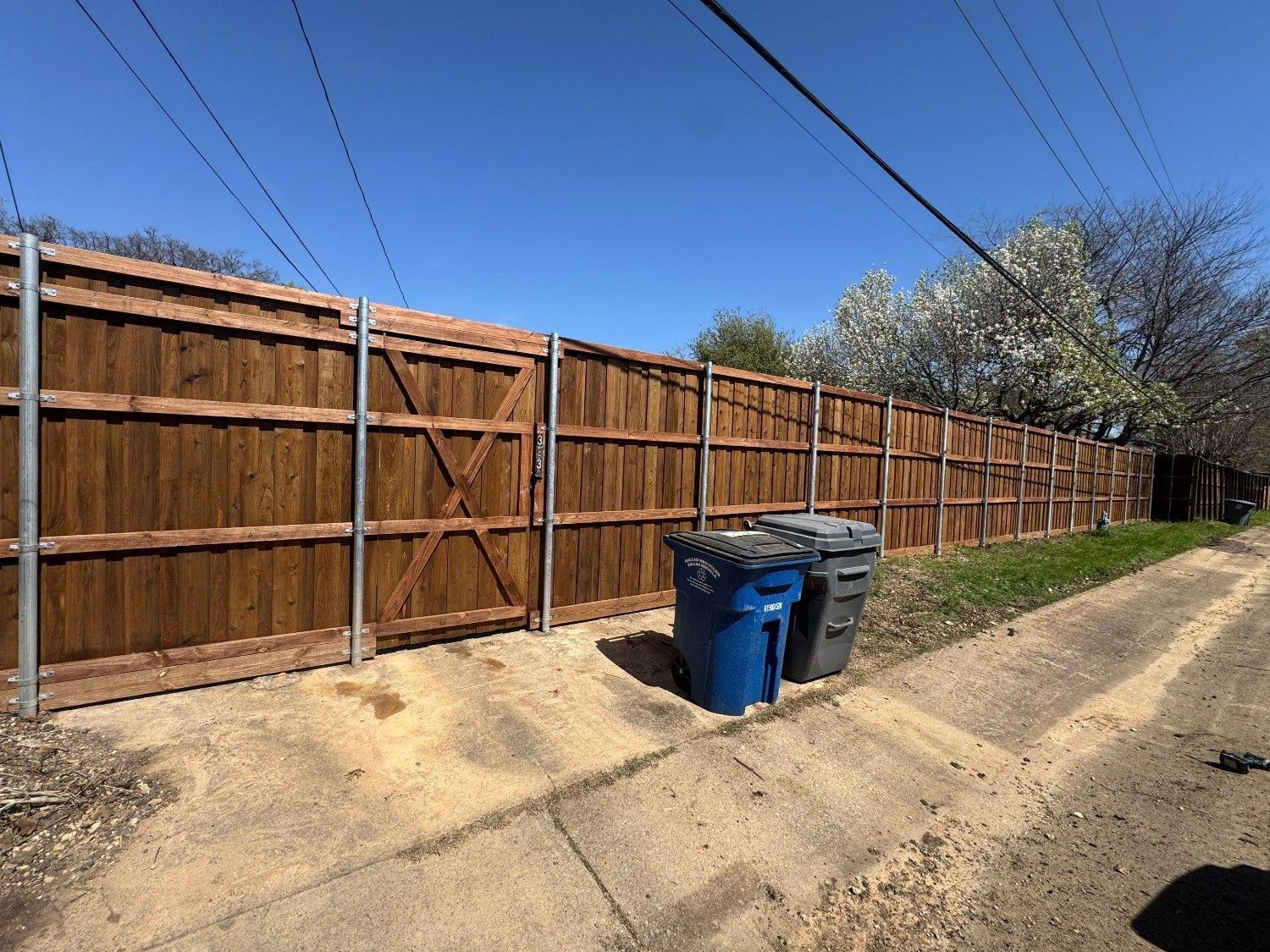 Long wooden fence beside a dirt alley with two trash bins under a clear blue sky.