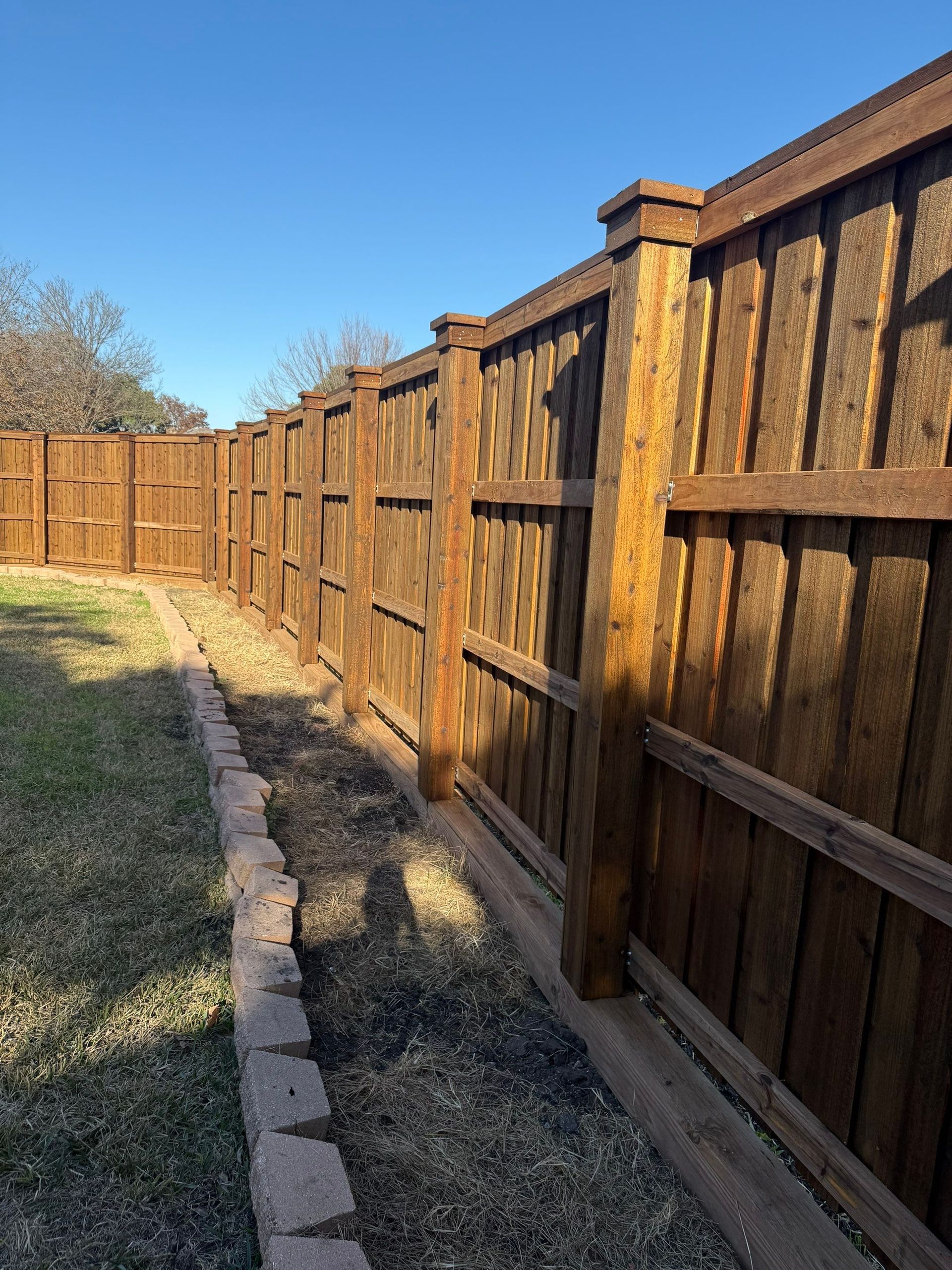Wooden privacy fence along a grassy yard beside a paved path under a clear blue sky