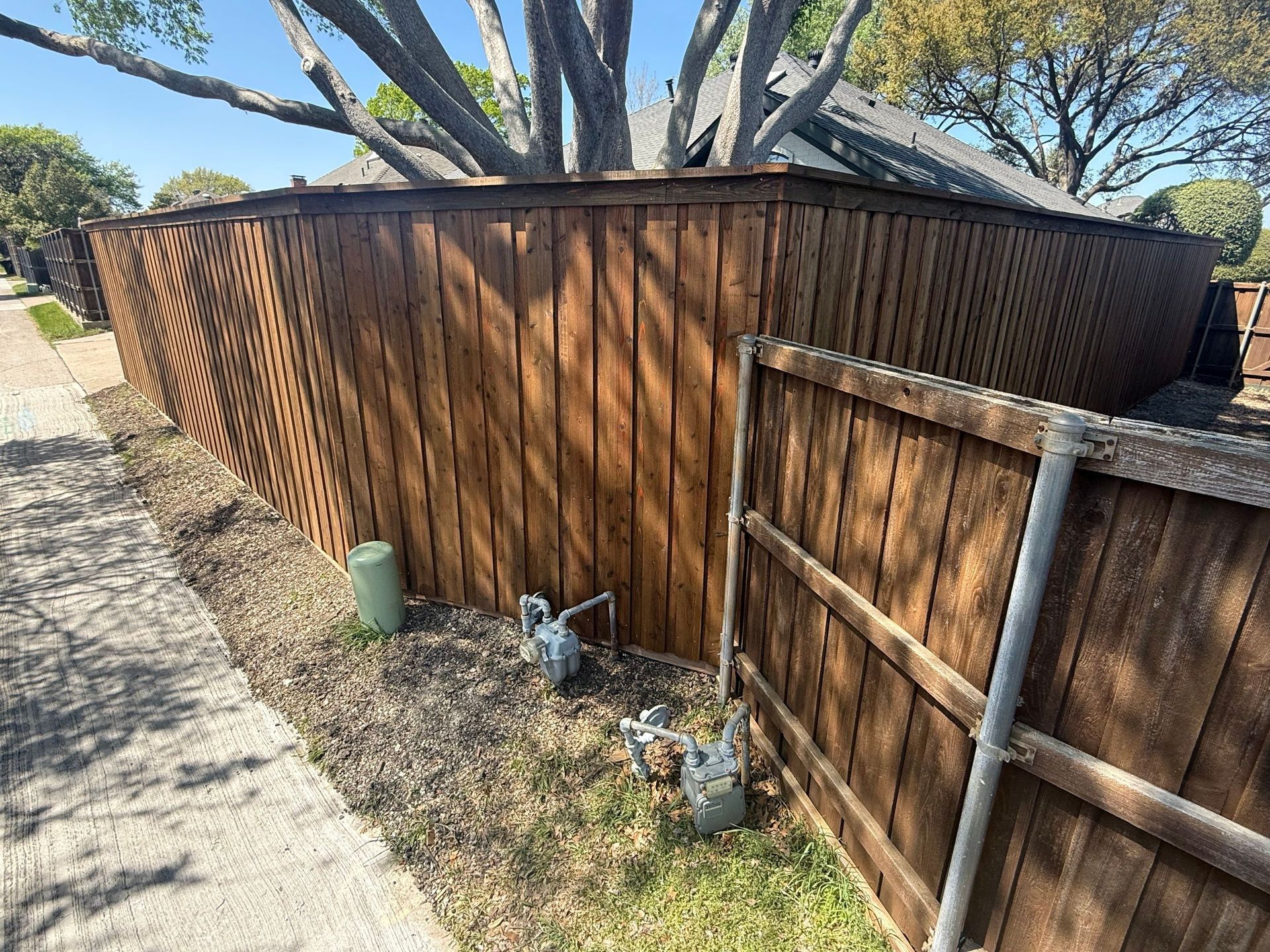 Wooden privacy fence and gate beside a sidewalk, with utility meters and trees overhead.