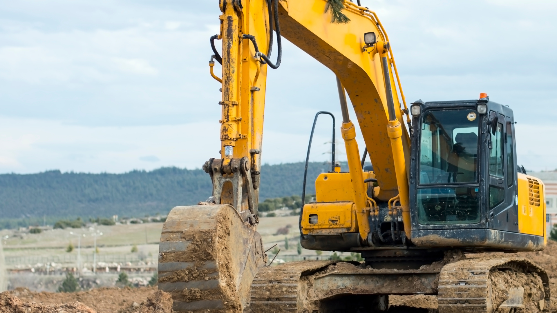 Yellow excavator on a dirt site with bucket raised, near water and hills in the background