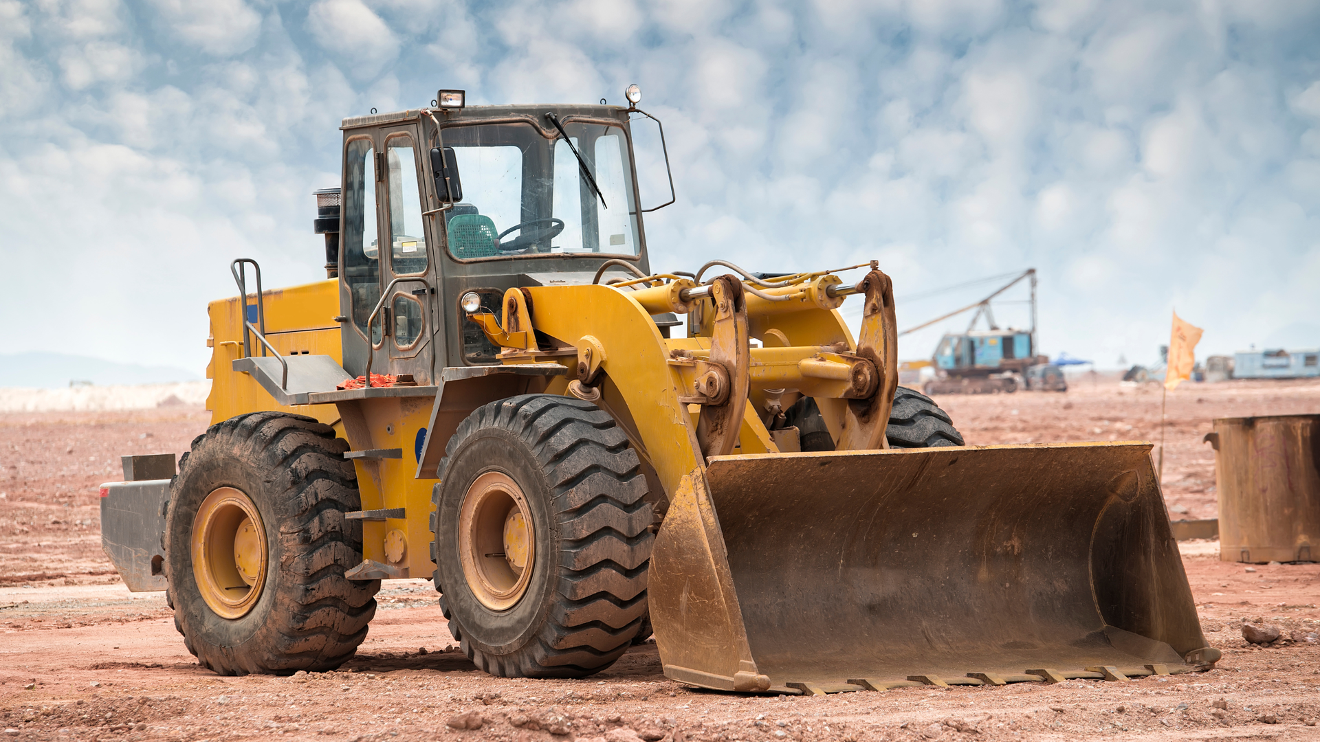 Yellow front loader on a construction site, holding a large dirt blade against a dusty ground.