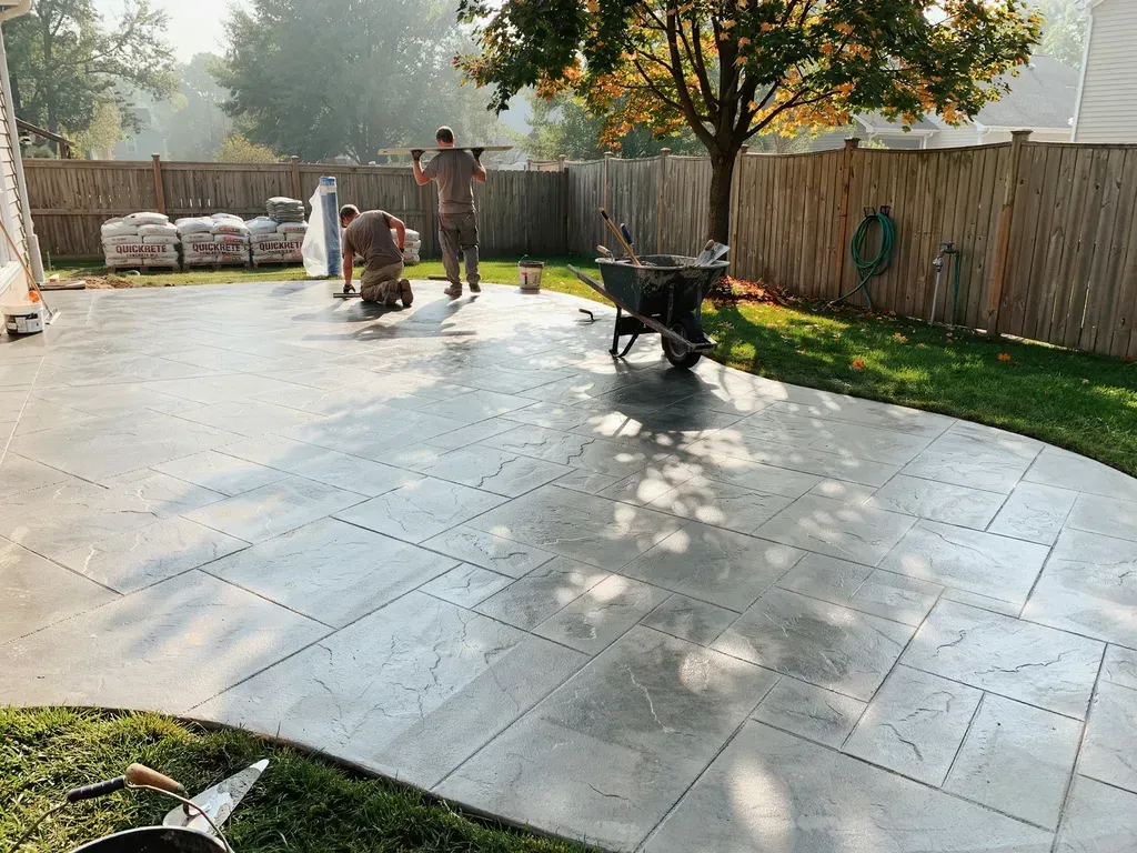 Workers pouring a large concrete patio in a fenced backyard, with a wheelbarrow and tree nearby.