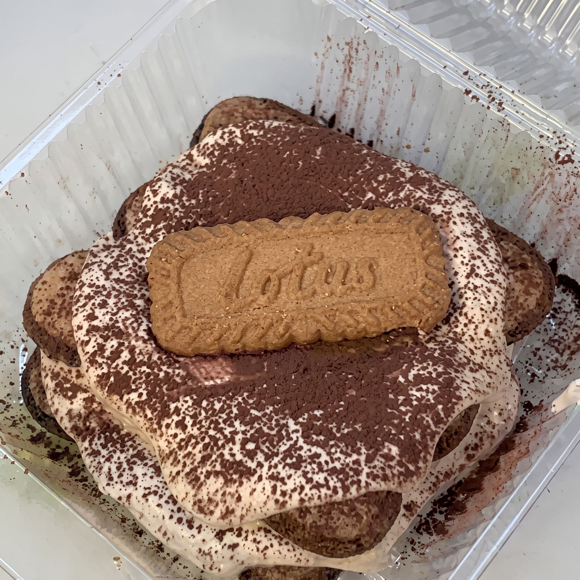 Tiramisu dessert in a clear plastic container topped with cocoa powder and a Lotus biscuit
