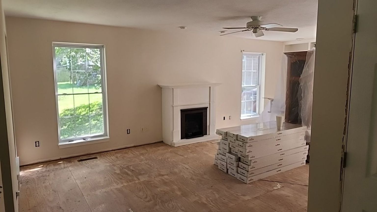 Living room under construction; bare wood floor, fireplace, two windows, and stacked materials.