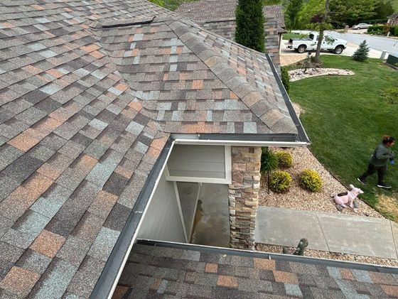 A house's asphalt shingle roof, dark gutters, and part of the porch, with a person in the yard.