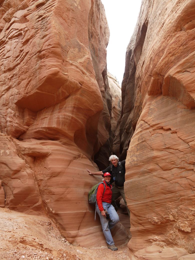 Two climbers in red and gray gear squeeze through a narrow red sandstone slot canyon.