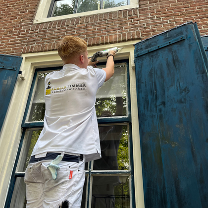 Schilder die een raam in een bakstenen gebouw met blauwe luiken afkit.