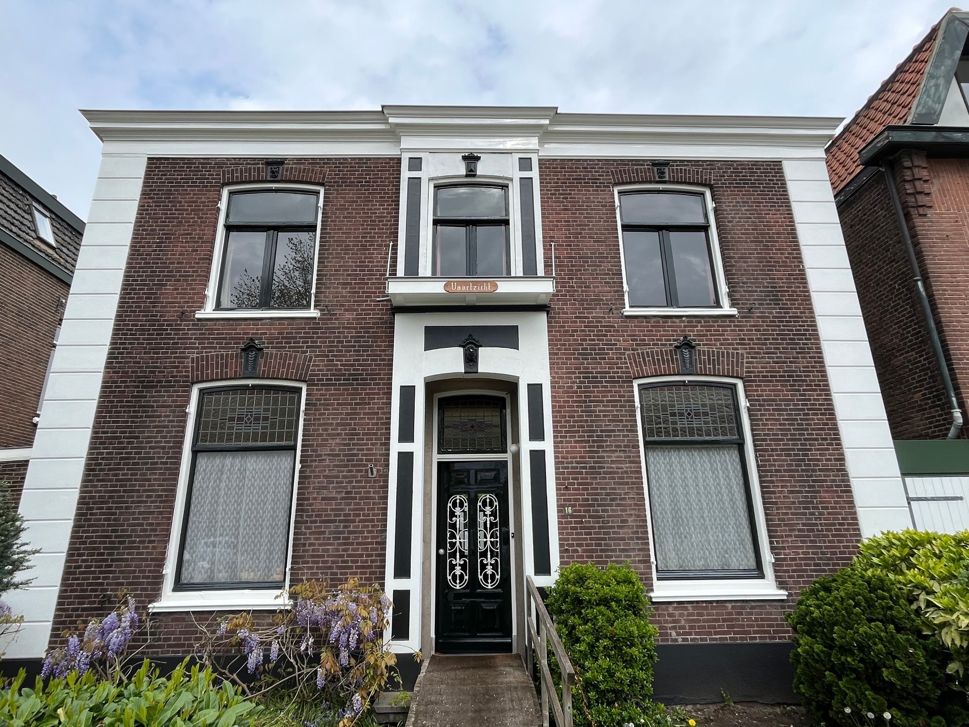 Bakstenen gebouw met zwarte ramen, deur en witte kozijnen. Struiken en groen aan de voorkant.