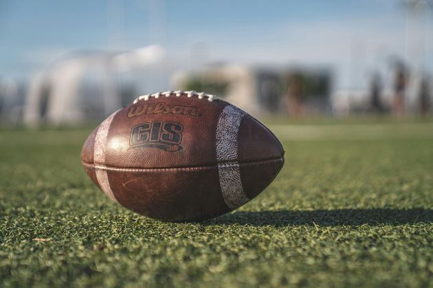 Flag football game at night, a player in red jersey runs with the ball as others pursue on a green field.