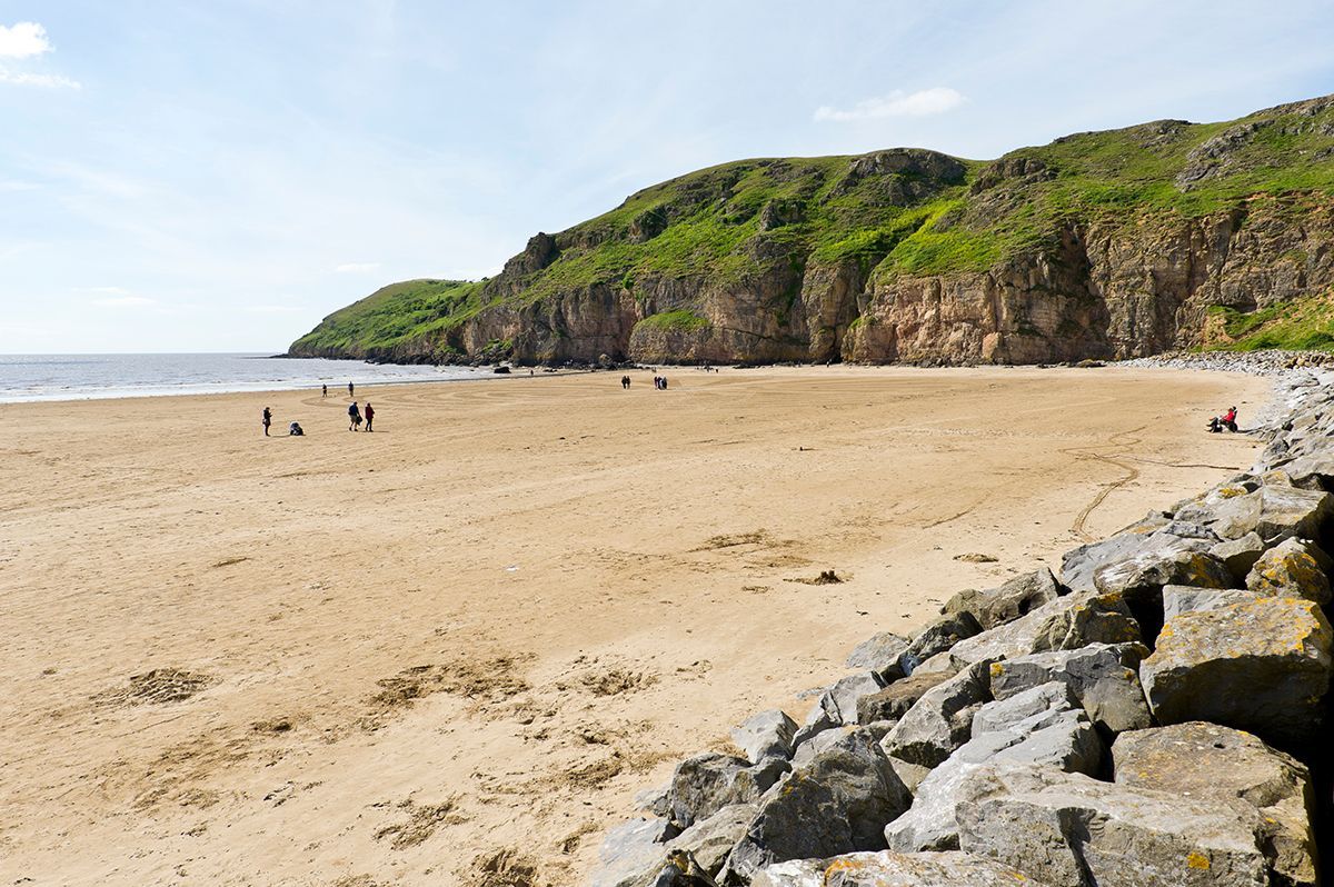 A group of people are walking on a beach next to a rock wall.