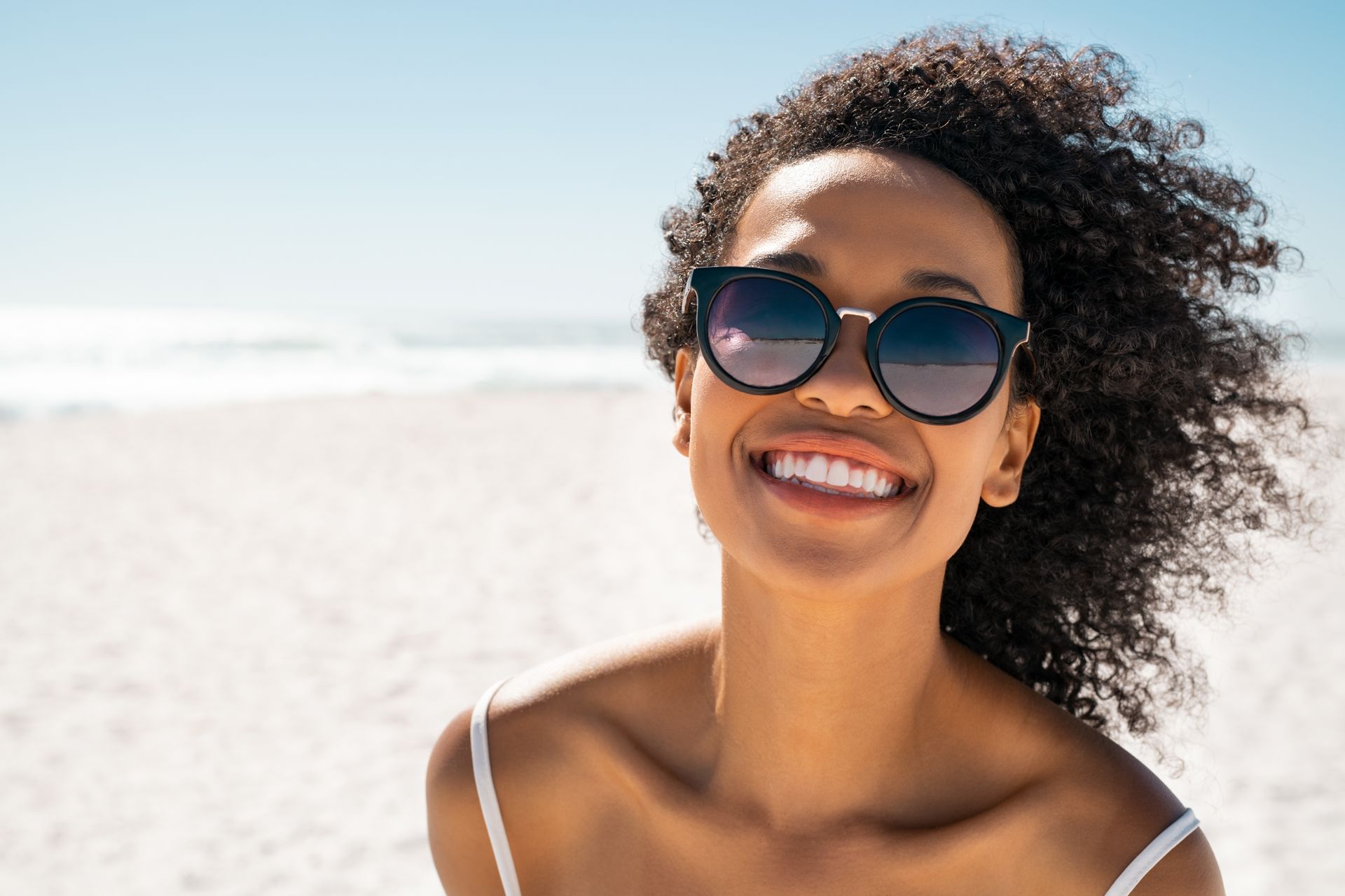 Person with curly hair standing on a sunny beach with the ocean in the background.